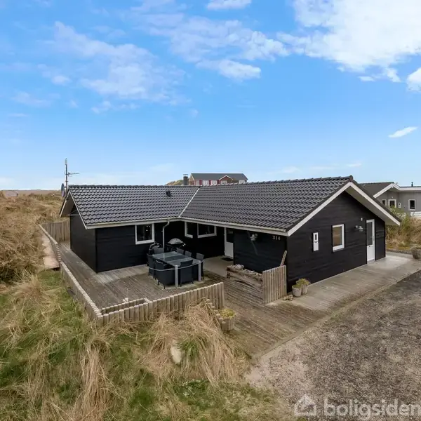 Et sort træhus ligger stille blandt klitter med en træterrasse, hvor et havemøbelsæt står. Huset er omgivet af græs og sand, under en klar blå himmel.