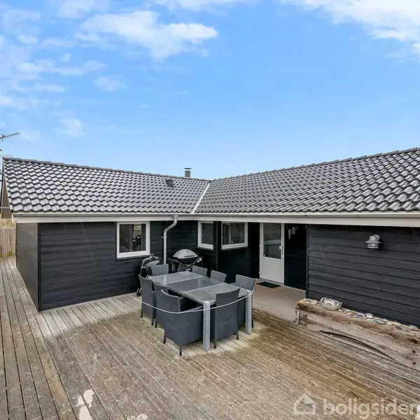 Stue med grå hjørnesofa og rundt bord, dekoreret med puder og stearinlys. Store vinduer giver udsigt til haven. Lyse trævægge og skråt loft skaber en hyggelig atmosfære.