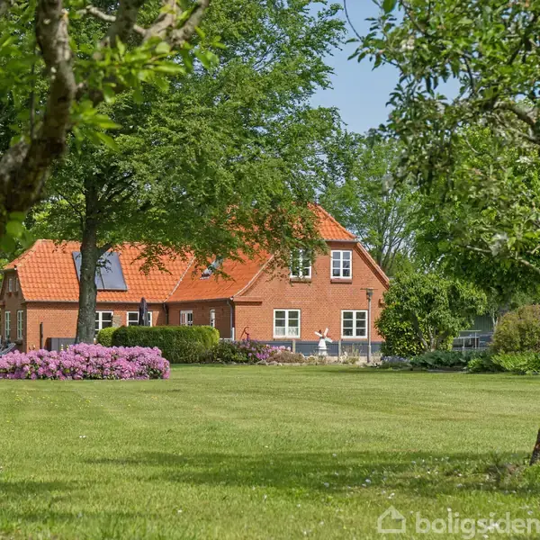 Rødt hus med tegltag står omgivet af grønne træer i en velplejet have med grøn græsplæne og blomsterbede.