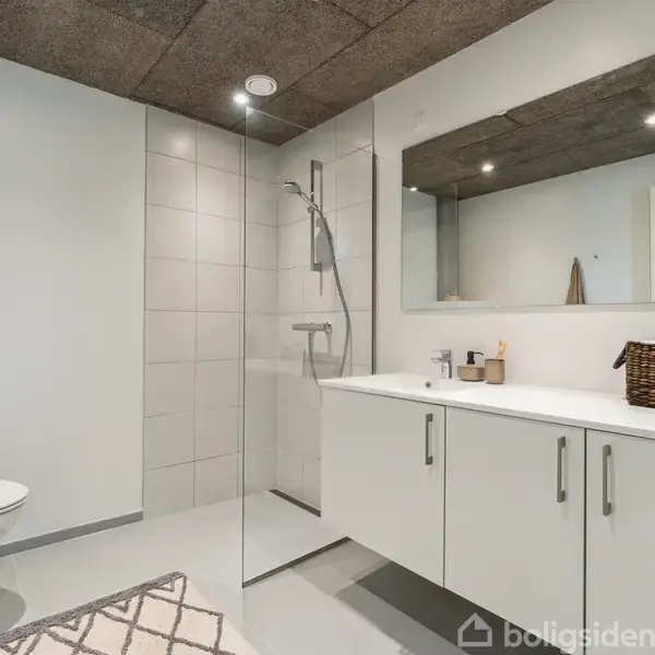 Et moderne badeværelse med væghængt toilet til venstre. En glasvæg adskiller bruseområdet. En hvid håndvask med skuffer og spejl hænger på højre væg. En kurv står på bordet.