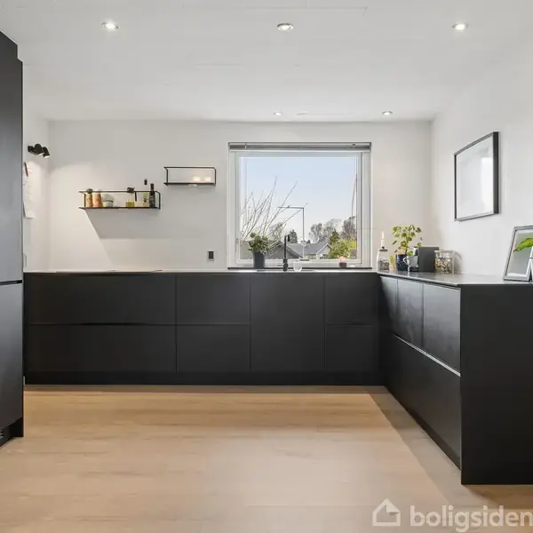 Moderne køkken med sorte skabe og indbyggede ovne; en stor, lys vindue skaber en venlig atmosfære. Hylder med krydderier og planter tilføjer dekoration. Trægulv understøtter det stilfulde look.