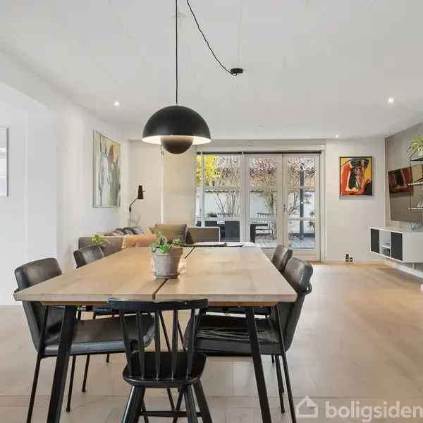 Spisebord med sorte stole er centralt placeret, omgivet af en moderne stue med sofa, kunstværker på væggene og store vinduer, der giver udsigt til en terrasse.