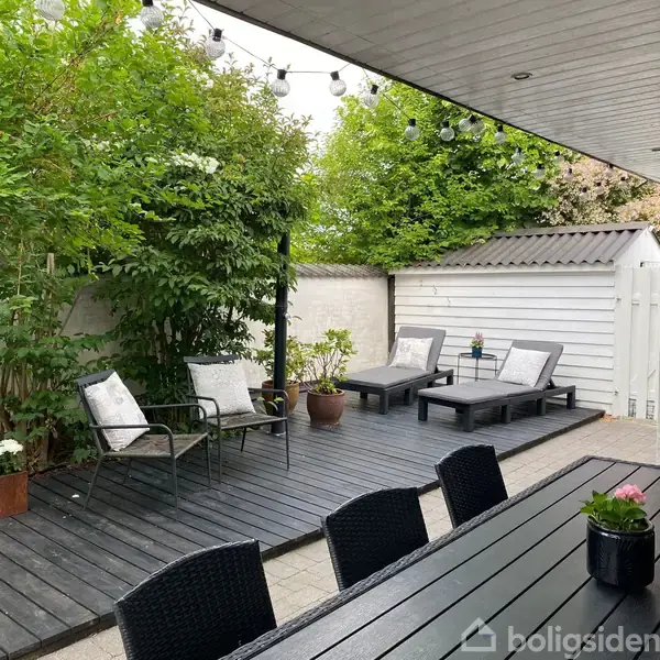 Et hvidt, én-etagers hus står på en grusbelagt grund med en parkeret bil foran. Huset har sorte tagsten, med omgivende planter og et skovområde i baggrunden.