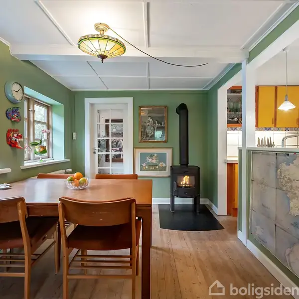 Spisebord med fire stole står i et grønt malet rum. En brændeovn opvarmer miljøet ved siden af dør med glasruder. I baggrunden ses køkkenet med orange skabsfronter.