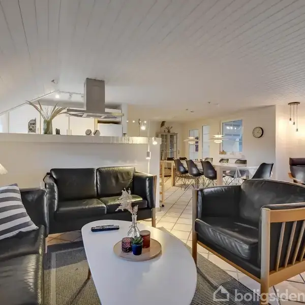 Sort lædermøblement med to sofaer og en stol står omkring et hvidt sofabord i en stue. Åben køkken og spisestue i baggrunden med lys klinkegulv og hvide vægge.