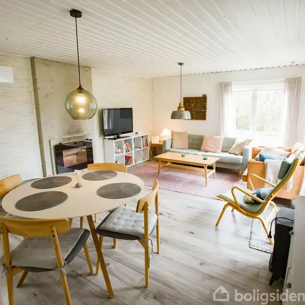 Rundt spisebord med fire stole står i et lyst, velindrettet stueområde. Sofa og sofabord står ved vindue. Lamper hænger fra loftet, og en reol med TV er i hjørnet.