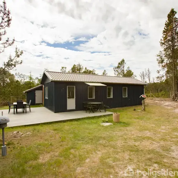 Et sort sommerhus står stille på en plæne med træer omkring. En terrasse med bord og stole ses i forgrunden under en overskyet himmel.