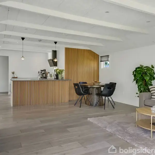 Stue med en moderne, lys indretning; en sofa står ved siden af et sofabord, mens spisebord og stole står nær køkkenø. Grønne planter og træelementer skaber en hyggelig atmosfære.