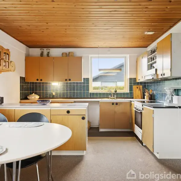 Et køkken med lysebrune skabe og hvide bordplader. En rund spisebord med stole står i forgrunden. Rummet har træloft og flisevægge. En vindue giver naturligt lys.