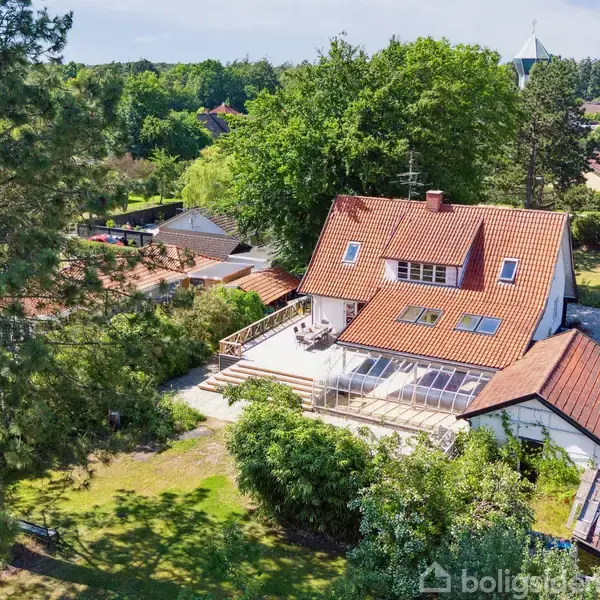Huset med rødt tegltag står i en grøn have, omgivet af træer. En stor terrasse er foran, og der er en sti til venstre i et boligområde.