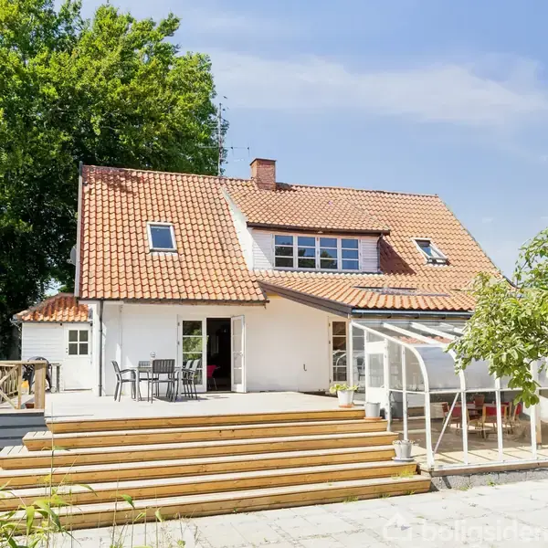 Huset har rødt tegltag og er omgivet af en stor træterrasse. Et bord med stole står på terrassen ved siden af en gennemsigtig overdækning. Baggrunden viser træer under en blå himmel.