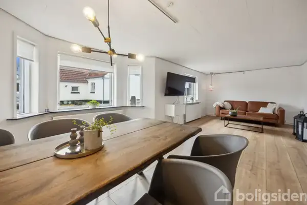 Træbord omgivet af grå stole står i spisestue med hængende lampe; stuen har en brun sofa, stort TV og store vinduer, der viser udsigt til taglejlighed udenfor.