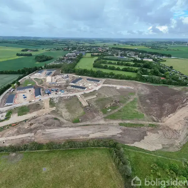En byggeplads under udvikling ligger centralt i landskabet omgivet af marker og skove. Bygninger er ved at blive konstrueret i den bagerste del, mens store jordområder er klargjort til yderligere arbejde.