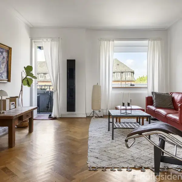 Rød sofa står i moderne stue med trægulv og kunst på væggene, omgivet af naturligt lys fra store vinduer. En lænestol og sofabord suppleres af planter på skrivebordet.
