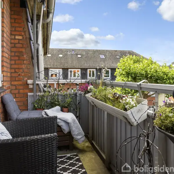 Fletstol og sofa står på en altan omgivet af planter og blomsterkasser. En tæppe pryder gulvet. Baggrunden viser en ældre murstensbygning og grønne træer under en blå himmel med skyer.