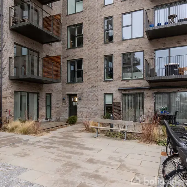 En moderne lejlighedsbygning med altaner står stille i et gårdmiljø med cykler og bænke. Bygningens facade er af mursten, og nummeret 19 vises ved indgangen.