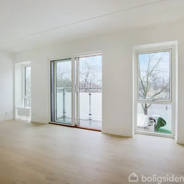Et lyst værelse med trægulv har store vinduer og en glasdør åben til en balkon. Udenfor ses træer og en hvid væg, der skaber en rolig atmosfære.