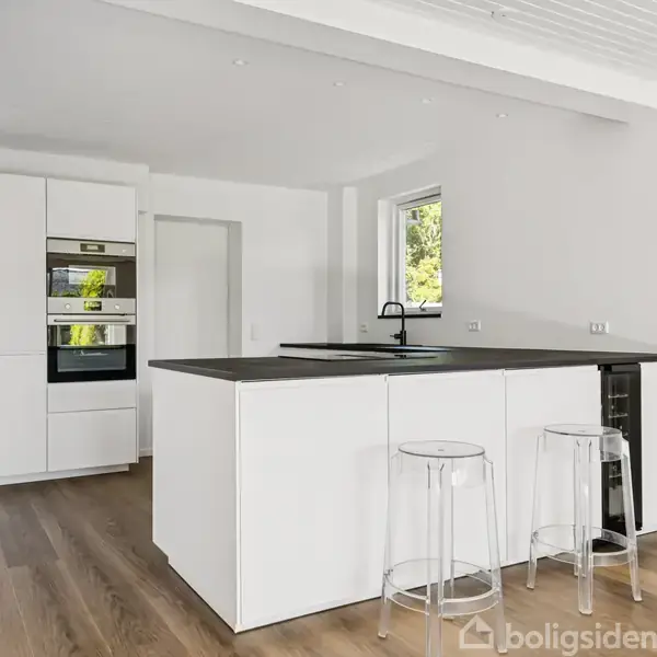 Køkken med hvidt køkkenø, sorte bordplader, to gennemsigtige barstole. Indbygget ovn og skab i baggrunden, trægulv og et vindue giver dagslys. Moderne og lyst design.