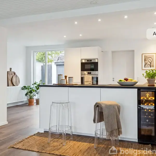 Køkken med hvid køkkenø, to gennemsigtige barstole og indbygningsovn i skinnende, moderne indretning. Planter og frugtskål pynter på bordpladen. Teksten "AI visualisering" ses øverst.