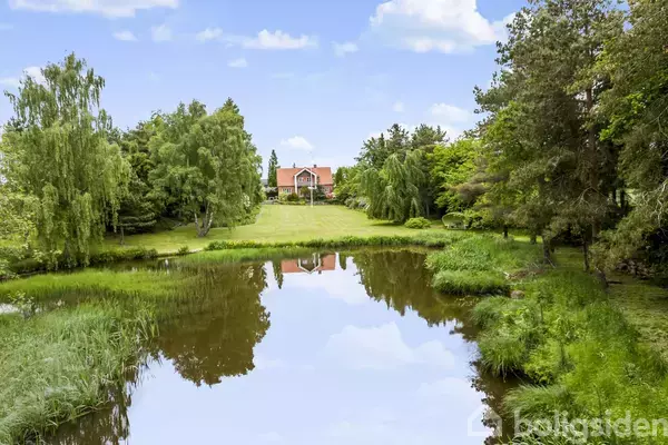 Et rødt hus står i baggrunden omgivet af frodige grønne træer, mens en rolig sø i forgrunden reflekterer himlen og omgivelserne.