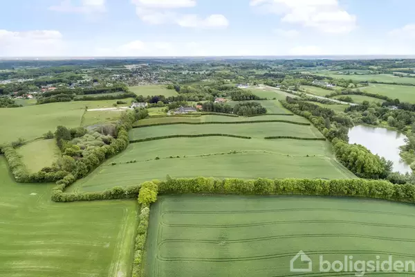 Landskabsudsigt viser grønne marker og en lille skov, med en sø til højre og spredte huse i det fjerne under en blå himmel med lette skyer.