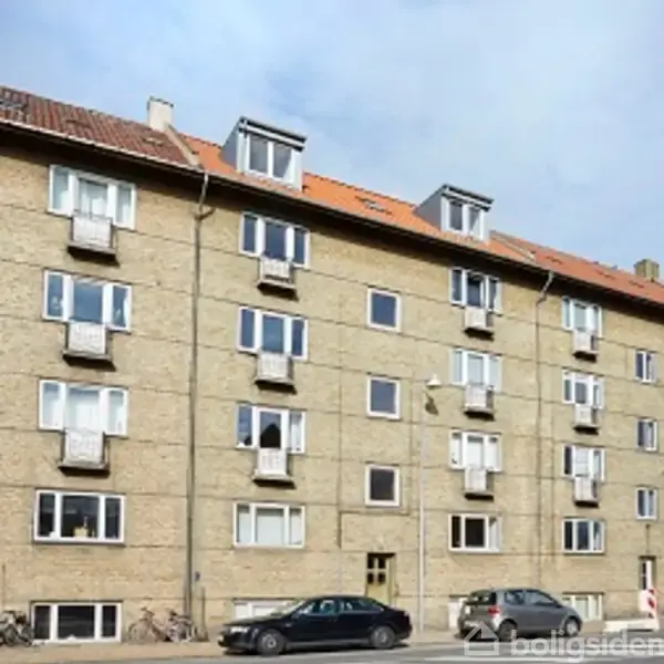 Flereetagers bygning med fyrretræsfacade har åbne vinduer og små altaner. Taget er dækket med røde tegl. Bygningen er placeret i et byområde under en blå himmel.