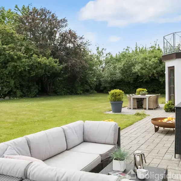 En udendørs sofa står på en terrasse i en have. Sofaen er omkranset af planter og et fliseområde. I baggrunden ses et bord, træer og en stor græsplæne.