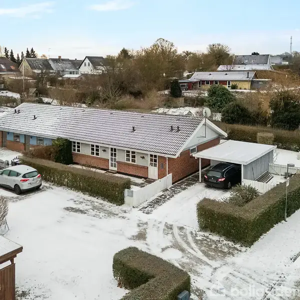 En lav, rød murstensbygning er omgivet af sneklædte områder med parkerede biler og en campingvogn foran. Hække omkranser ejendommen i et boligkvarter med flere huse i baggrunden.
