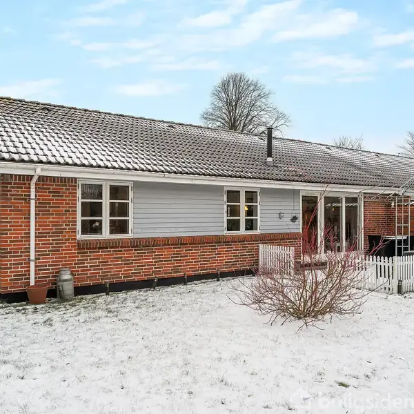 Et rødt murstenshus med hvide vinduer står på en snedækket grund. Huset har en satellitantenne på taget og en lille veranda med et hvidt stakit foran.