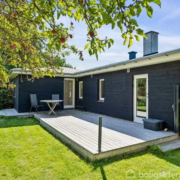 Et sortmalet træhus med en træterrasse og havemøbler står omgivet af en grøn græsplæne og træer med modne æbler hængende fra grenene.