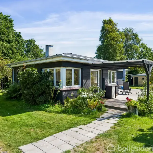 Et lille, træbeklædt hus med store vinduer står i en frodig have. En flisebelagt sti fører til indgangen, omgivet af grønne buske og træer under en klar blå himmel.