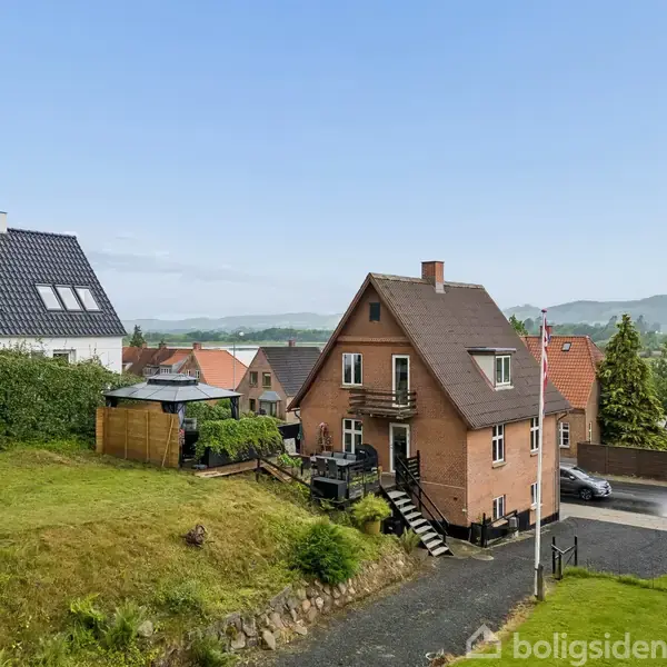 Et brunt murstenshus med stejl trappe, beliggende i en bakket boligområde. Haven er grøn med et lysthus i nærheden, og i baggrunden ses skovklædte bakker og flere huse.