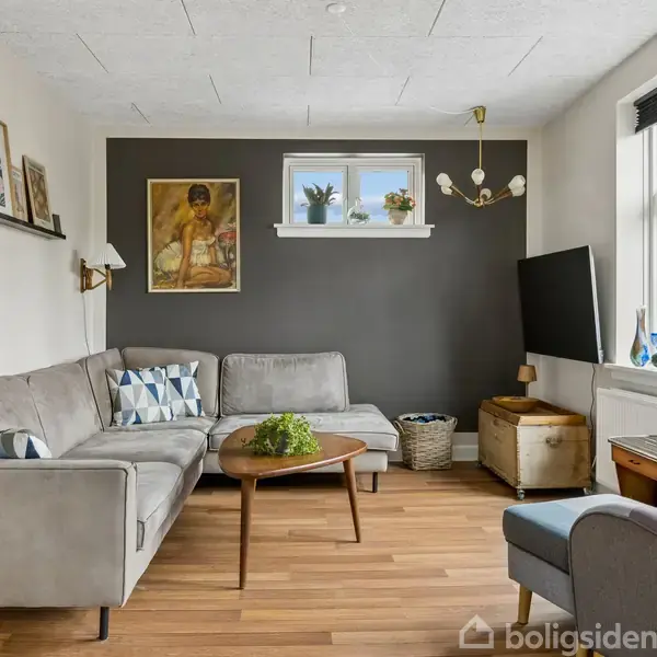 Grå sofa står i en stue med trælaminatgulv. På væggen hænger malerier, og et lille vindue lader lys ind. En lænestol og tv er placeret ved vinduet.