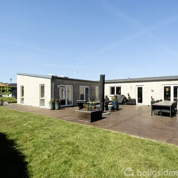 Hus med fladt tag og hvid facade står på en solrig terrasse. Omkring terrassen er en græsplæne med grøn hæk. Blå himmel i baggrunden.