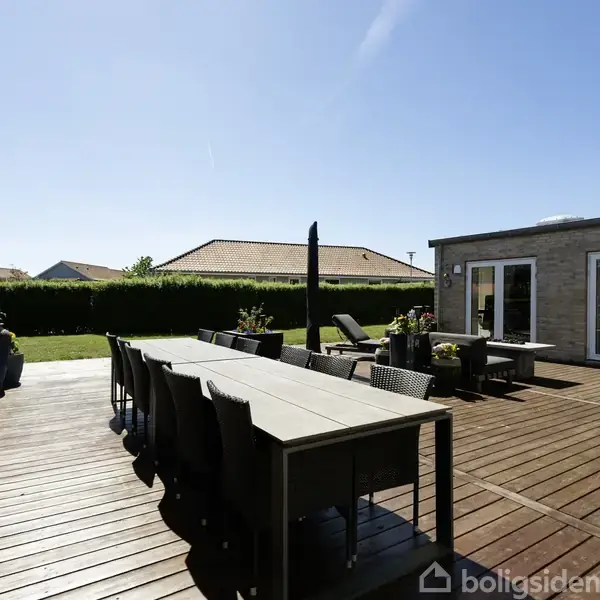 Terrasse med moderne havemøbler står klar til brug i solskin. Flere siddepladser og parasol omgivet af husfacade og grøn hæk i baggrunden.