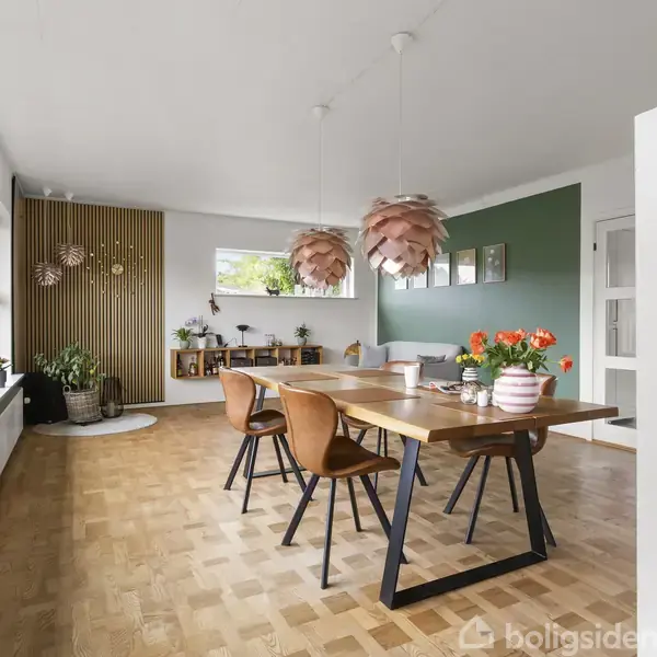Et spisebord med træstole står i et lyst rum med parketgulv. To lyserøde lamper hænger over bordet. En grøn væg har billeder, og der er dekorationer med blomster.