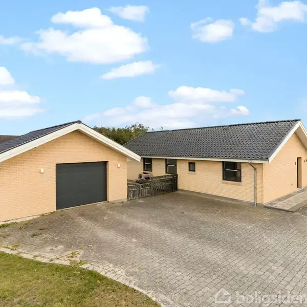 Et lysegult murstenshus med sort tag står i et åbent område. Foran huset er en flisebelagt indkørsel og en garage. Blå himmel med skyer i baggrunden.