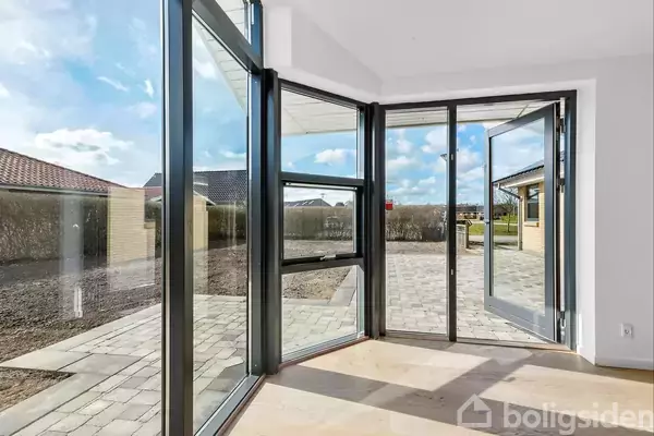 Glassdøre åbner ud til en stenbelagt terrasse i en have. Dørene reflekterer sollys, mens himlen er delvist overskyet. Terrasse omgivet af hæk og bygninger i det fjerne.