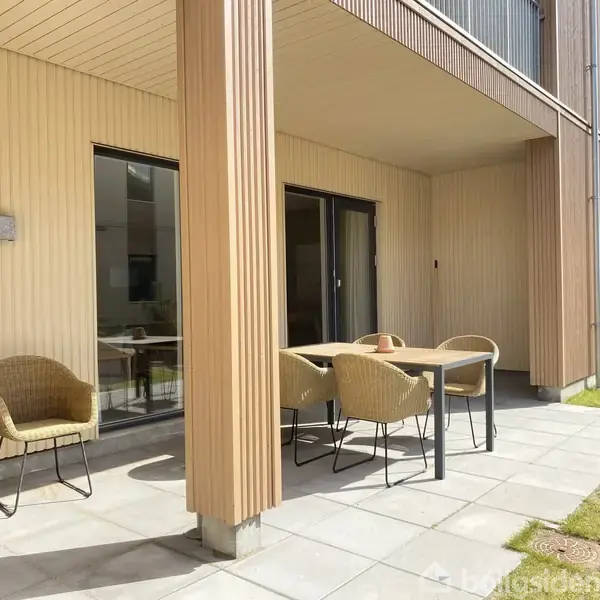 Terrassebord med kurvestole står på en flisebelagt terrasse foran et moderne byggehus. Skydeglasdøre bagved giver adgang til indendørsområder. Grøn græsplæne omgiver terrassen, og solen skinner.