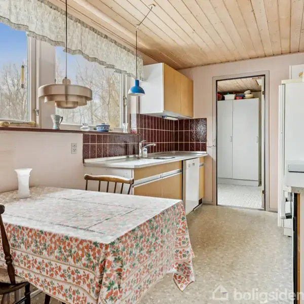 Et køkkenbord med blomstret dug står ved vinduet. Bordet har to stole omkring det. Køkkenet har brune fliser og træloft, med en håndvask og diverse apparater i baggrunden.