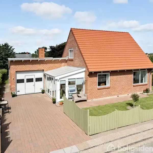 En murstensvilla med rødt tegltag og integreret garage står på en flisebelagt indkørsel. En lille, grøn have er omkranset af et lysgrønt hegn. Klar himmel og grønne planter omkring.