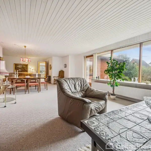 En lys stue har et gråt lænestol foran store vinduer, der giver udsigt til haven. Til venstre står en reol og et spisebord. En grøn plante tilføjer farve.