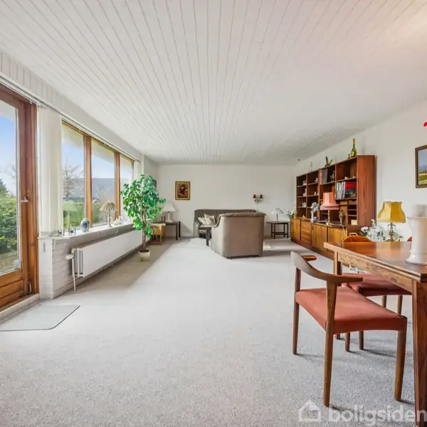 En stue med sofagruppe i baggrunden, spisebord med stole i forgrunden. Store vinduer lader lys ind. Reol, plante, og lysekrone skaber en hyggelig atmosfære i rummet.