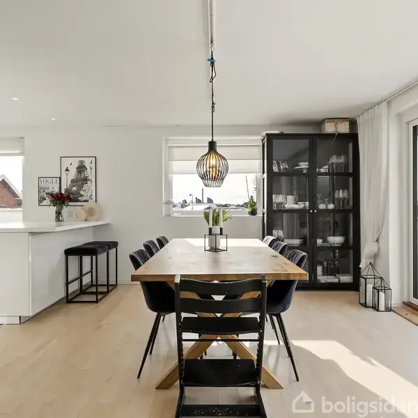 Bord står i centrum, omgivet af sorte stole i spisestue. Over bordet hænger en lampe. Til venstre er køkken, til højre store vinduer med udsigt. Indretningen er moderne.
