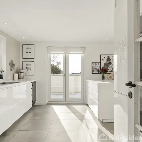 Lyst, moderne køkken med hvide skabe og bordplader. Franske døre leder til en balkon med udsigt. Væggene er dekoreret med rammer og kunst. Gulvet er flisebelagt.