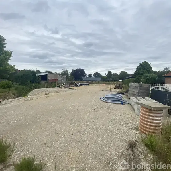 En grusbelagt byggeplads med byggeartikler spredt omkring i et åbent område. Træer og en rød bygning ses i baggrunden under en overskyet himmel.