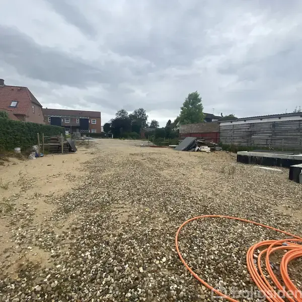 Grusbelagt gårdsplads med orange slanger til venstre, omgivet af huse og et træhegn, under en overskyet himmel.
