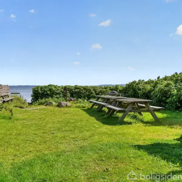 Et picnicbord står på en græsplæne ved vandet, omgivet af hække og buske. I baggrunden ses en træbænk og en sø. Himmel med få skyer.