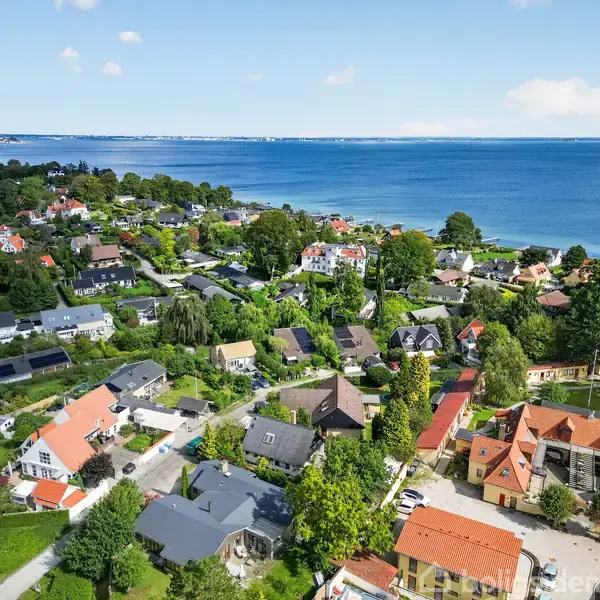 Huse står tæt i et nabolag med grønne træer; baggrund viser en vidstrakt kystlinje og blåt hav under en klar himmel.