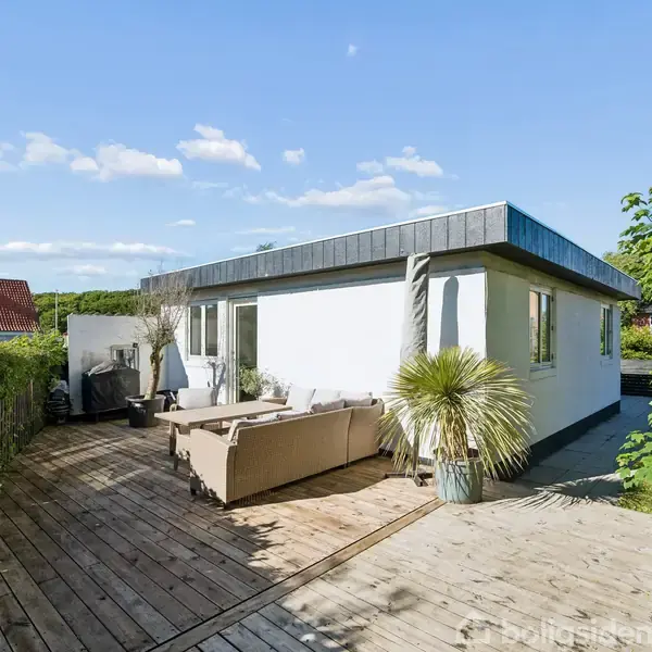 En hvid, moderne bungalow omgivet af en træterrasse med sofa og planter. Solbeskinnet dag i en stille forstad med grønne buske og blå himmel som baggrund.